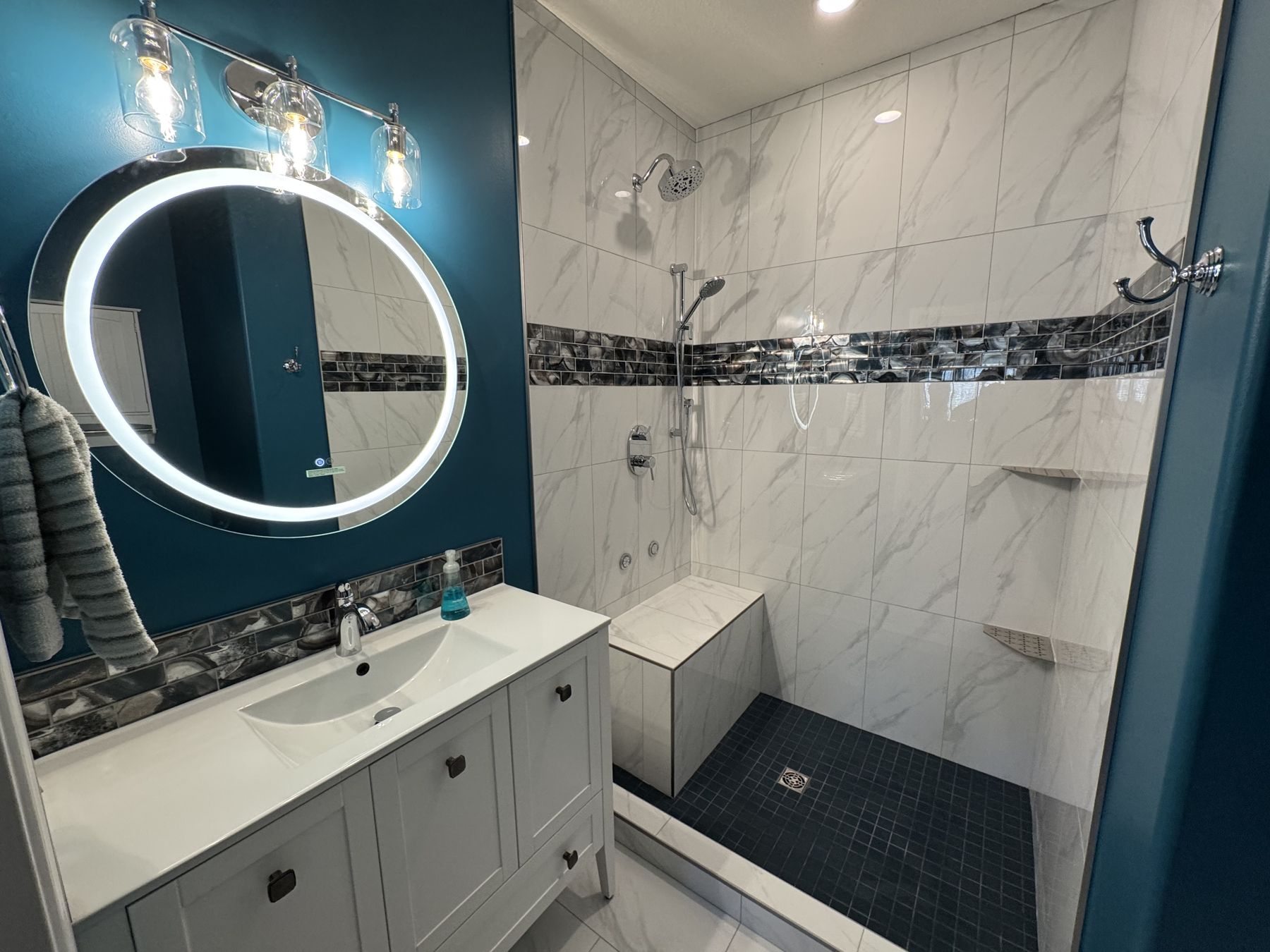 Renovated bathroom in Cochrane with deep teal walls, backlit round mirror, marble-look tile shower with mosaic accent band and bench