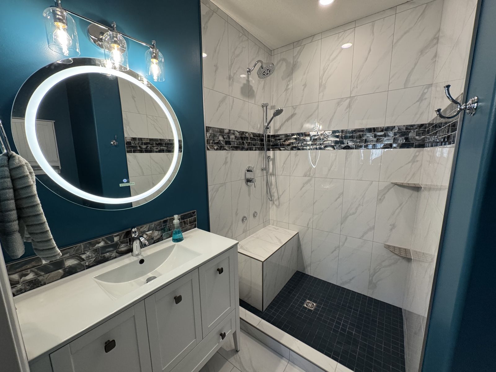 Bathroom with deep teal walls, backlit round mirror and marble-look tile shower