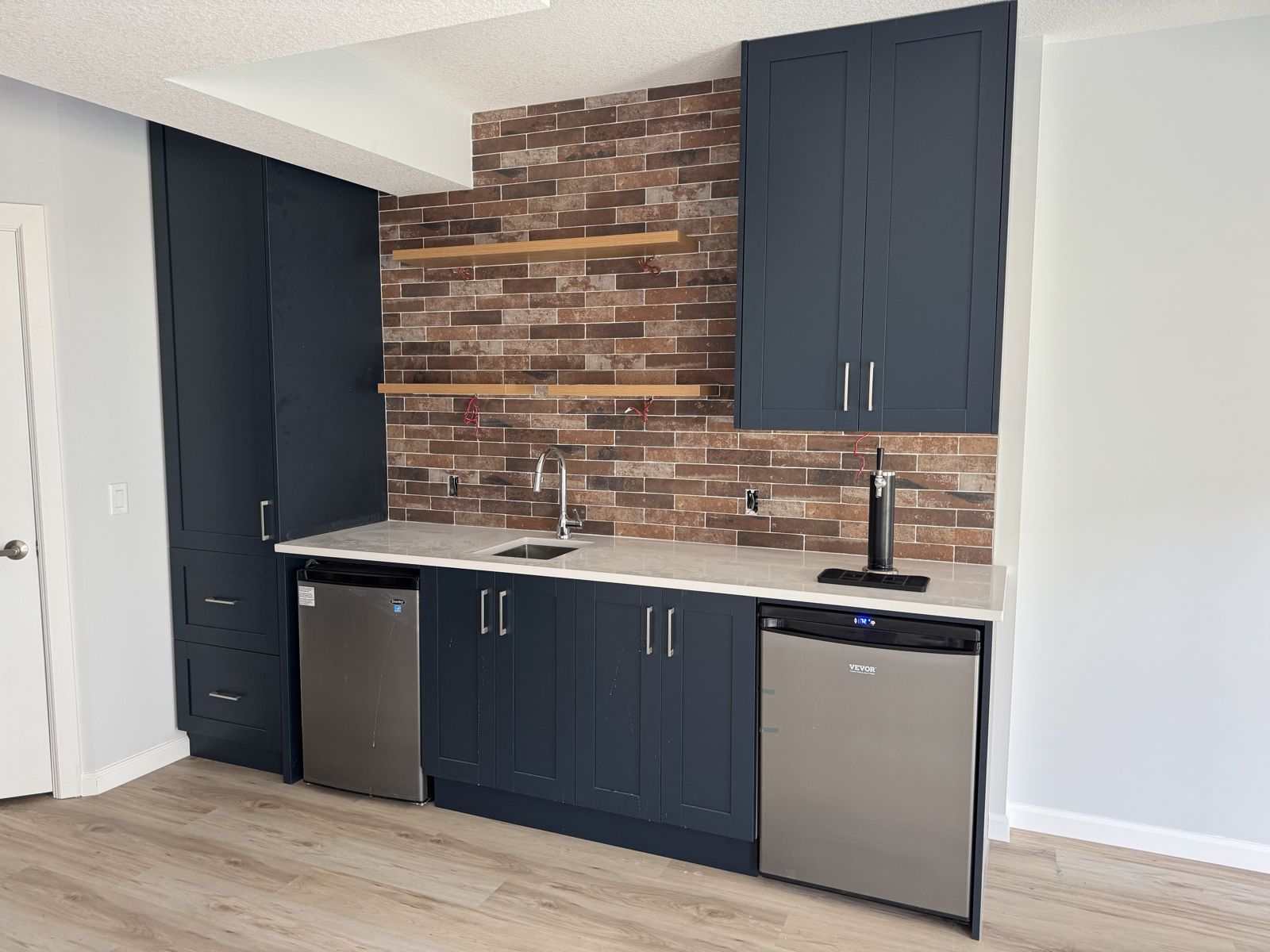 Finished basement wet bar with navy cabinets and brick backsplash